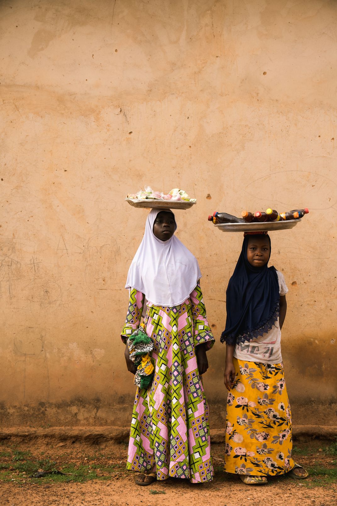 2 young African girls stand with trays on their heads