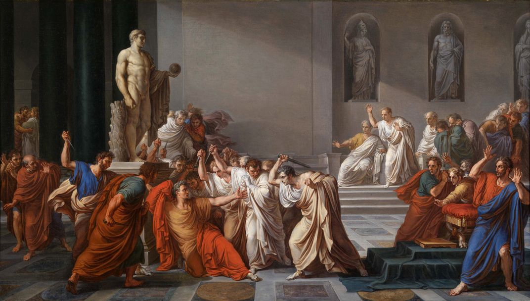 A Roman hall, filled with people in blue and red robes, people in white are attacking Caesar
