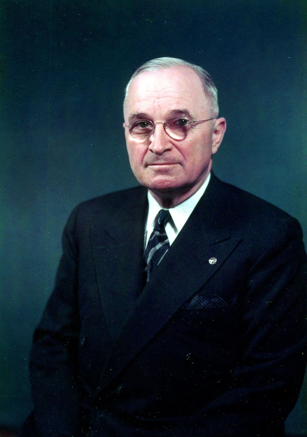 A color photograph of Truman, a white middle-aged man with gray combed hair, glasses, a serious expression, wearing a suit and tie