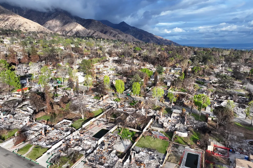 structures and rubble on lots with trees and grass in front of mountains