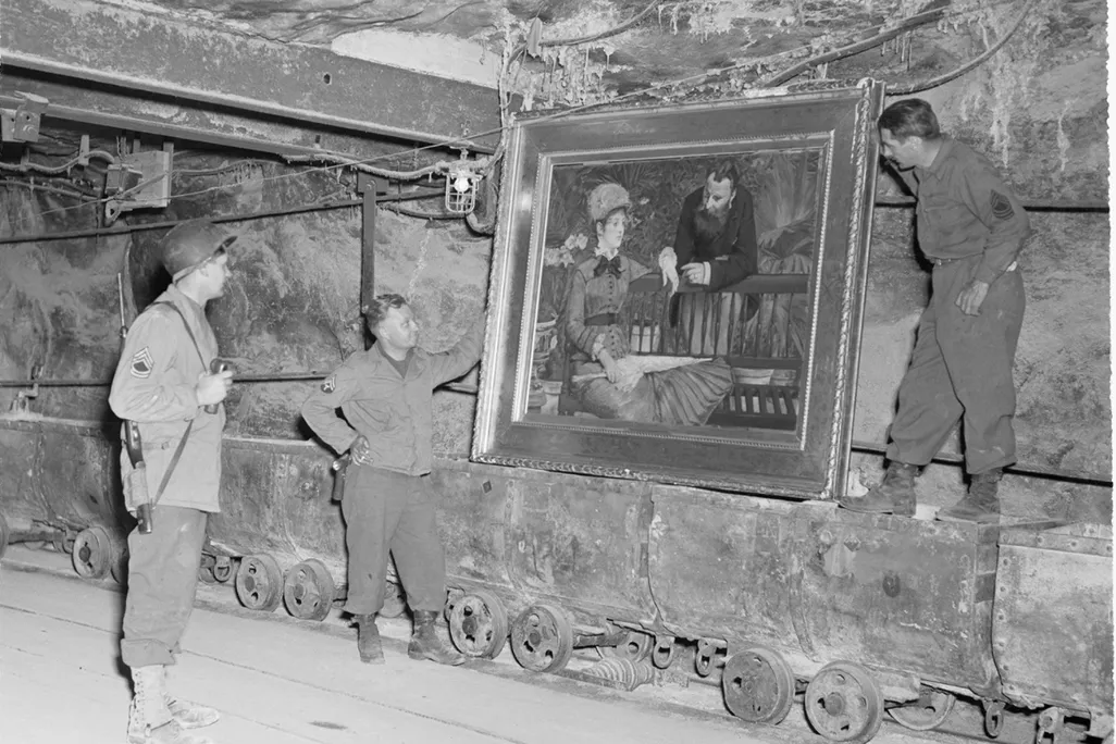 Three men in army gear stand in a bunker, holding a large painting of a man and a woman in an elegant frame
