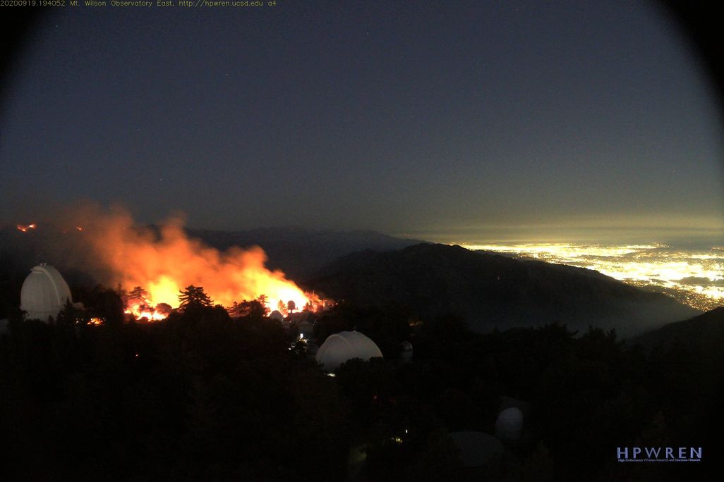 A panoramic shot of the soft glow of Pasadena and Los Angeles to the right, an expanse of dark nighttime hills, two white domed telescopes on the left with tall, bright orange and yellow flames bursting nearby, dramatic against the darkness