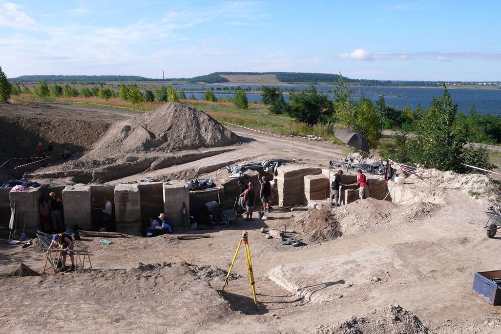 An image of archaeologists excavating a site. They stand in a pit near a mound of dirt. The surrounding area has lakes and trees.