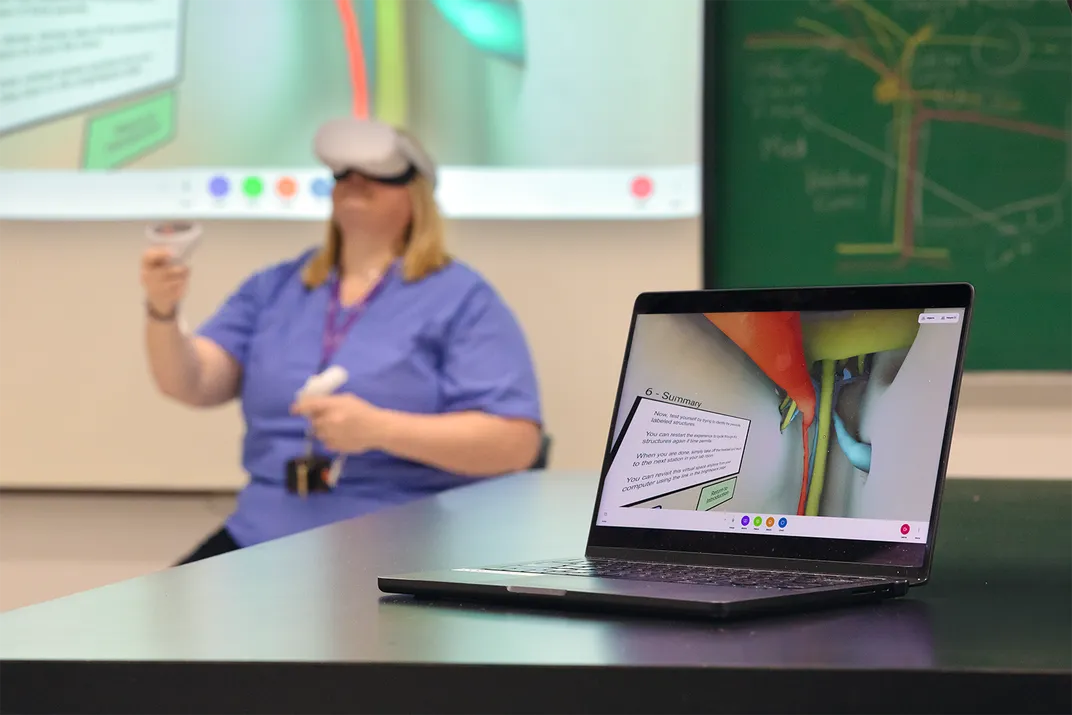 a computer shows graphics while a person sits in the background with a VR headset on