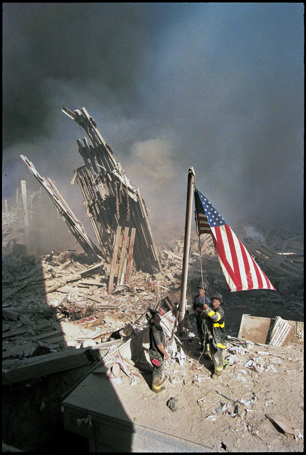 A vertical view of the scene, where the flag's bright red and blue colors stand out brightly against the yellow of the firefighters striped jackets and the muted smoke behind