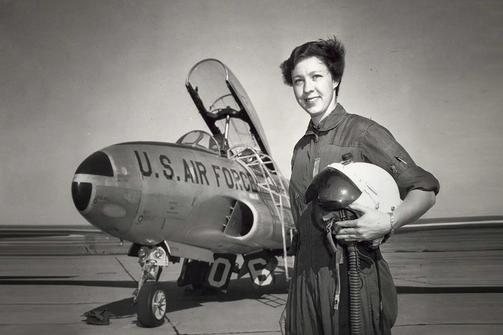 A photograph of a woman dressed to fly a jet, holding a helmet. The jet behind her has the top open and is labeled "U.S. Air Force"