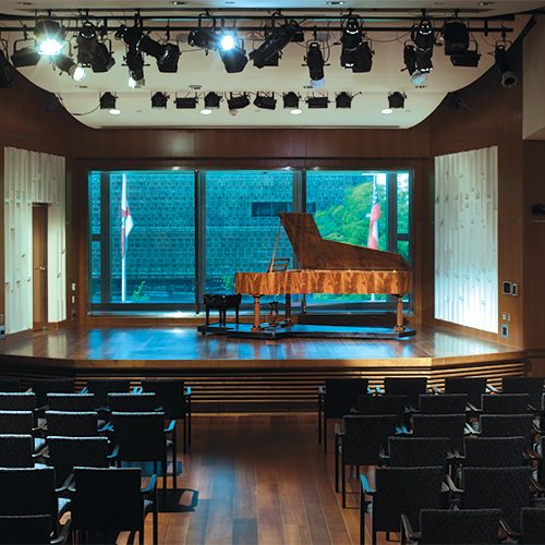 A grand piano sits on a stage in front of several rows of chairs