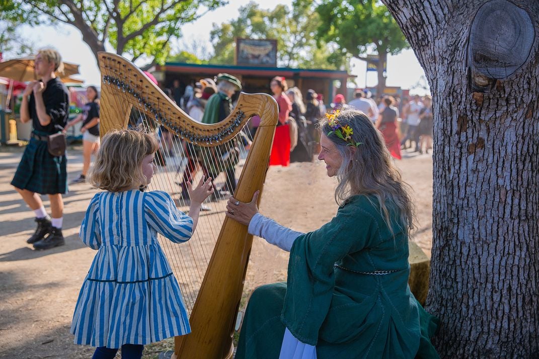 A young girl strums the strings of a harp held by her mother at an outdoor festival.