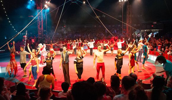 Performers wave, forming a ring around a red, circular stage, with a packed audience and rigging hanging above.