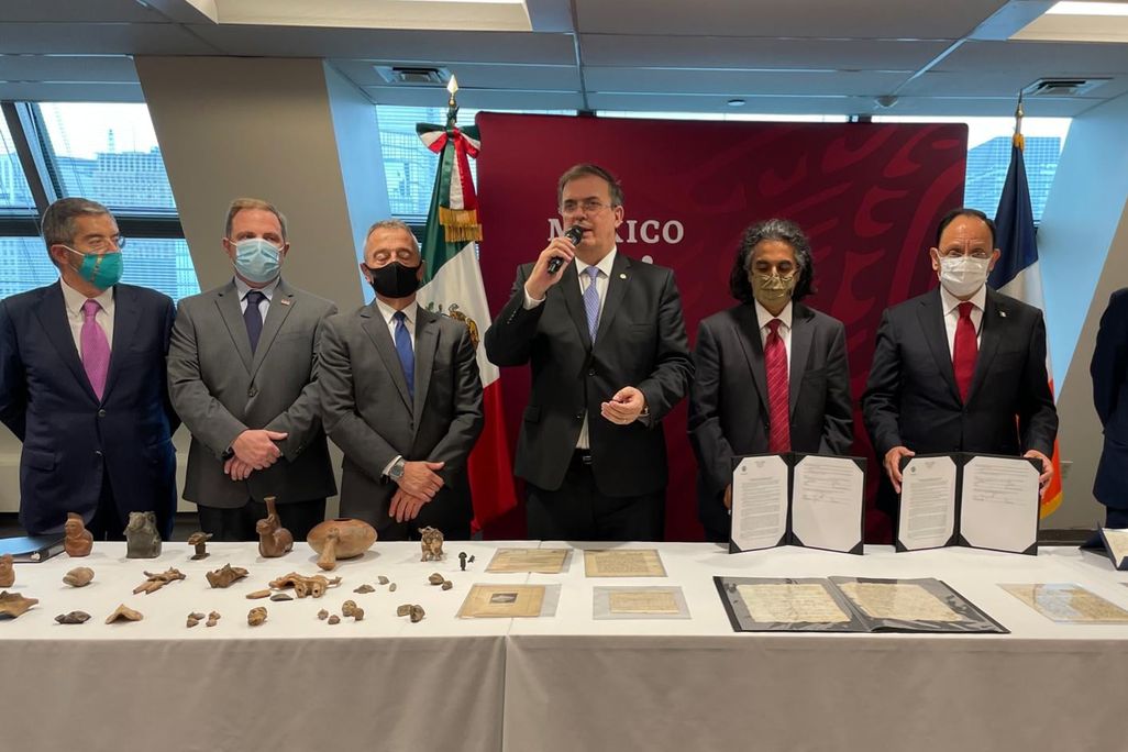 Seven people in suits and ties stand in front of a table with antiquities and 16th century documents laid out