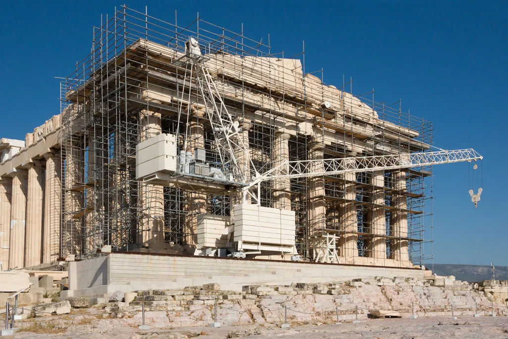 A view of the side of the Parthenon, on a sunny day with clear blue sky behind; the facade is covered in scaffolding and construction equipment