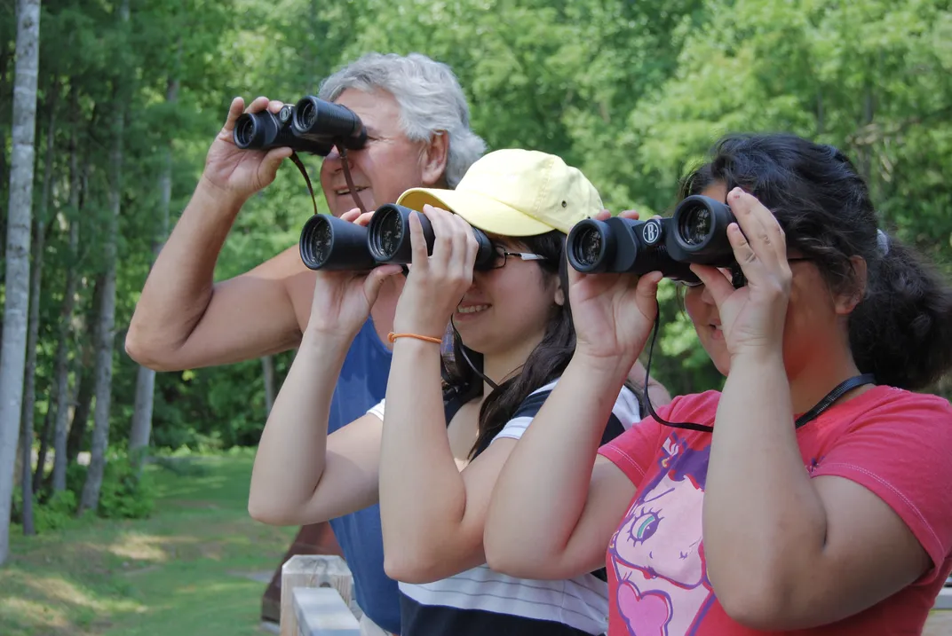 An older man and two younger women smile as they peer through binoculars along a forest boardwalk.