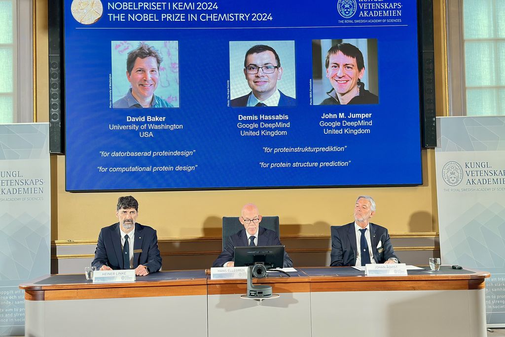 a slide showing the three Nobel winners is projected above three men sitting at a table