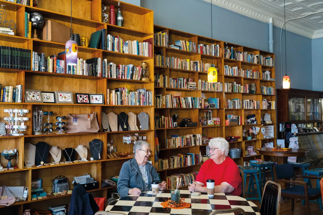two women sit chatting at a table in front of floor to ceiling bookshelves