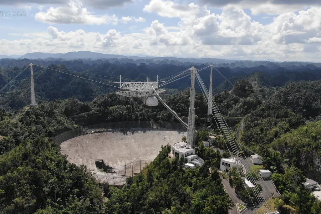 A photograph of the dish from high above. It has three tall towers around the circular dish, all connected by thick cables. The dish has a 100-foot hole on the side. The observatory is surrounded by expansive forest. 