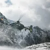 A helicopter flies low over a snow-covered, jagged mountain range. The rotors kick up a light mist of snow against the dark, rocky terrain.