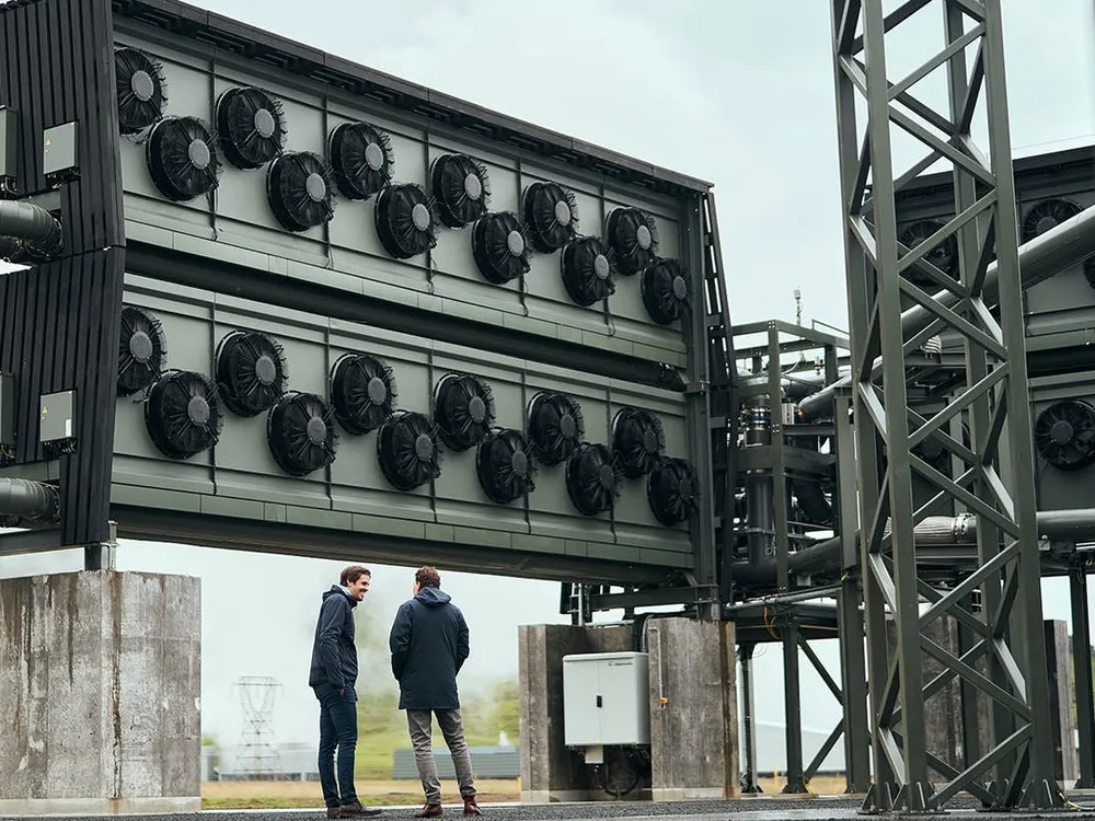 Two men stand in front of tall wall of fans at the Orca carbon capture facility in Iceland