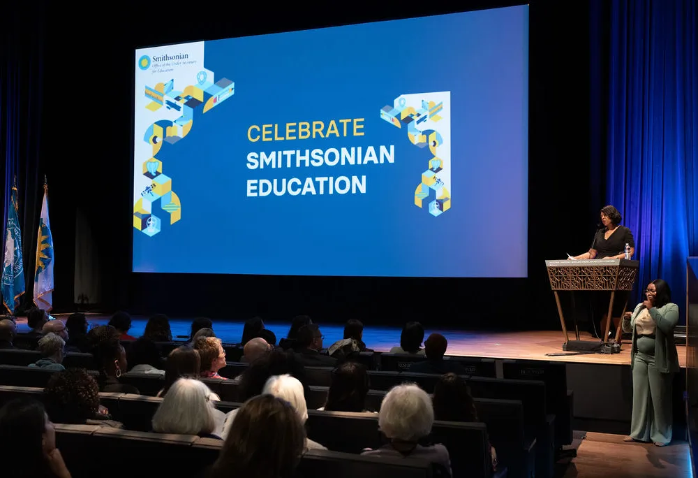 A woman stands at a podium in a theater with "Celebrate Smithsonian Education" projected on the screen