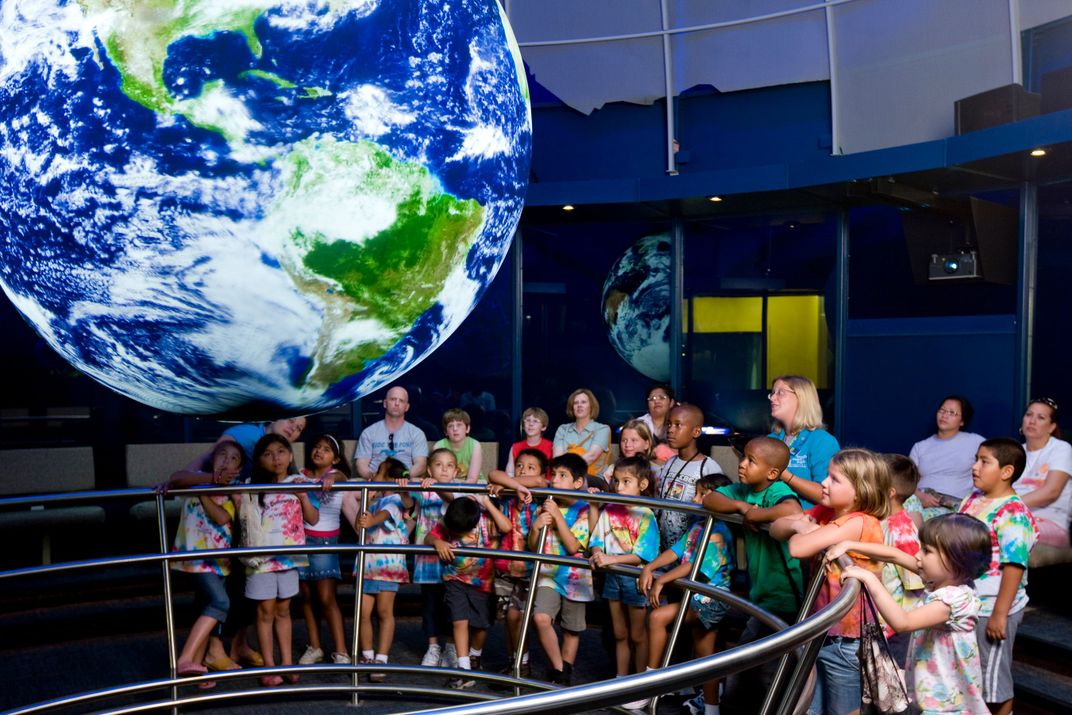 A group of children and teachers look up in awe at a seemingly floating, glowing reproduction of Earth.