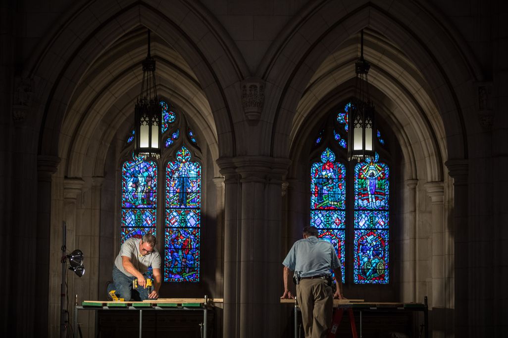 Workers erect scaffolding near two tall stained glass windows, which shine with blue light