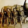 A black-and-white photo of four white women in their late 20s to early 30s walking on a military airfield. The women are wearing flightsuits, boots, and leather flight jackets. Behind the women is the forward fuselage of a World War II military bomber.