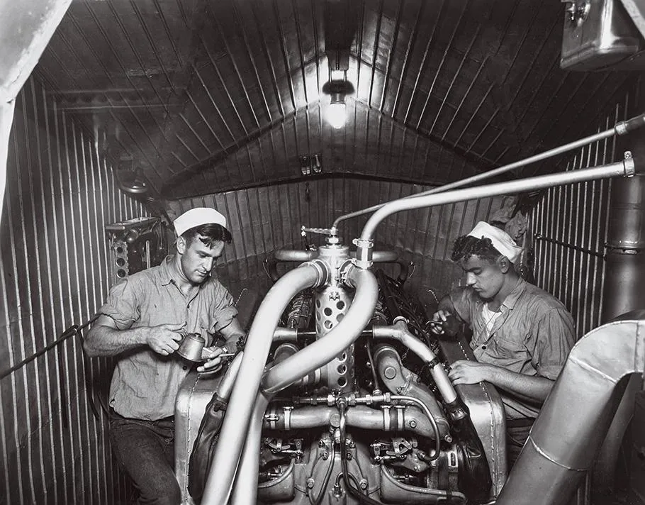 A black-and-white photo shows two Navy men, their sleeves rolled up wearing white hats, working on a device covered with several tubes of varying sizes.
