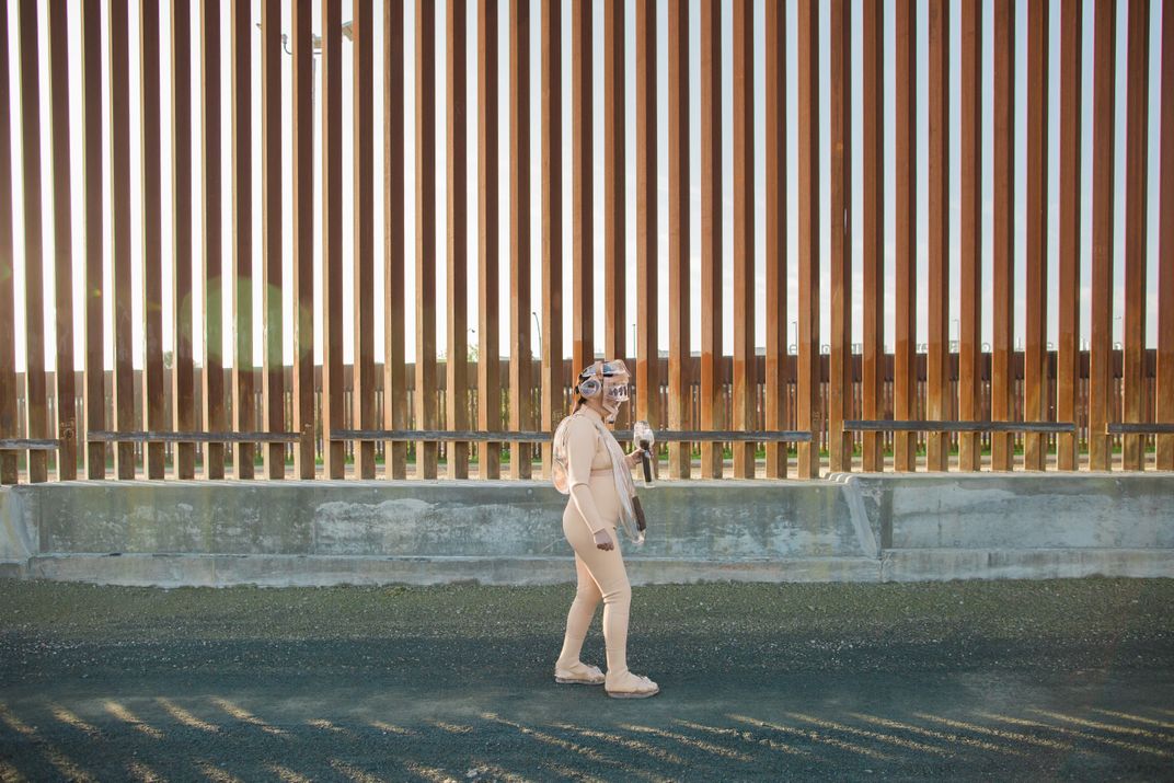 Artist Tanya Aguiniga walking in a suit made of various glass elements next to the U.S./ Mexico border wall.