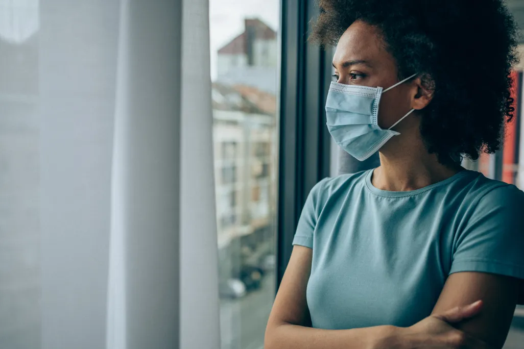 A Black woman with a face masks looks out of a window