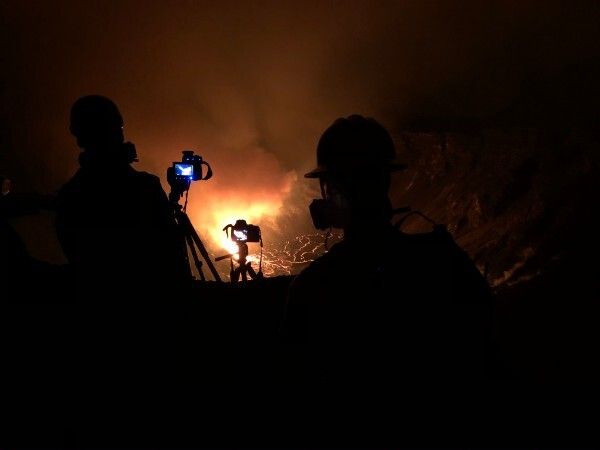 An image of the erupting volcano. Two people stand in the foreground with cameras, but only their silhouettes are visible. In the background, an orange cloud of steam is rising and the ground has bright orange crack in it. 