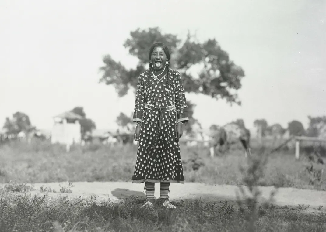 Black and white archival photo of Sarah Grandmother's Knife, age 10, in an elk tooth dress in a field playfully sticking her tongue out at the camera