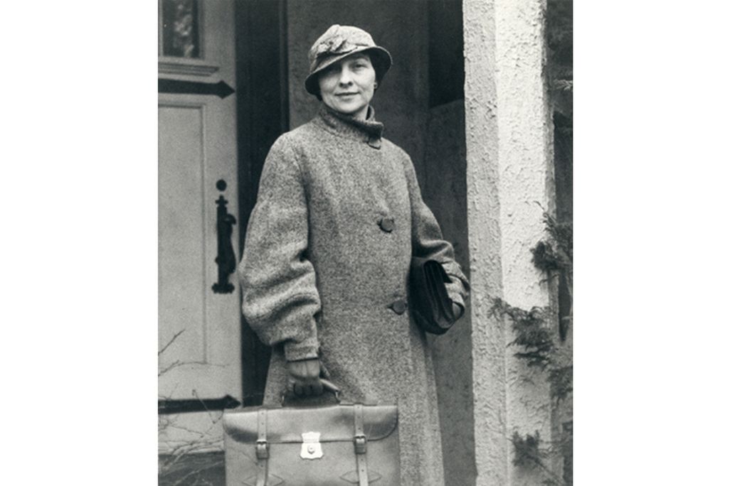 A young white woman in a long coat, wearing a fashionable hat cocked to one side on her head, carries a briefcase and poses in front of a doorway