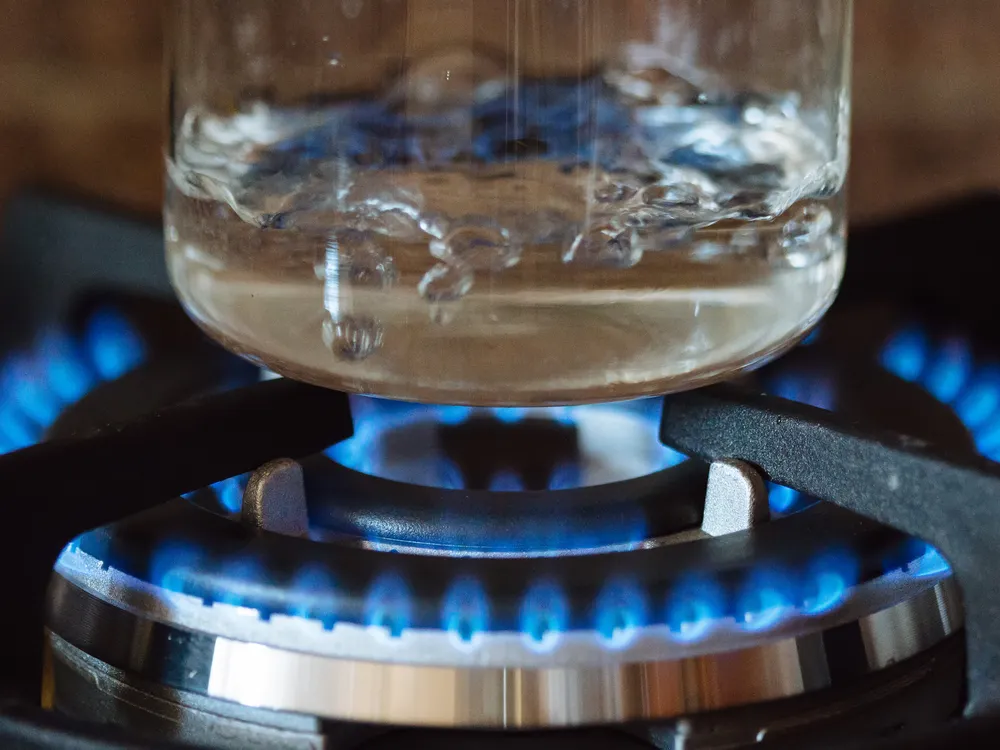 A close-up of a blue-flame burner on a stove, with water boiling in a clear glass pot.