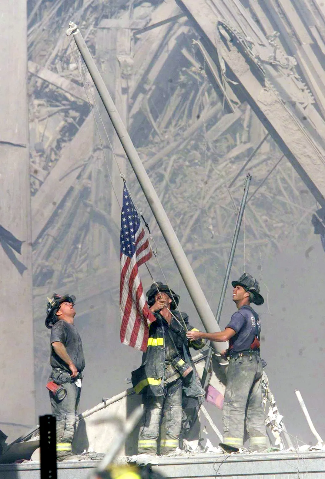 A vertical shot of three white men in the process of raising an American flag on a titled flagpole, which juts out of the rubble of Ground Zero