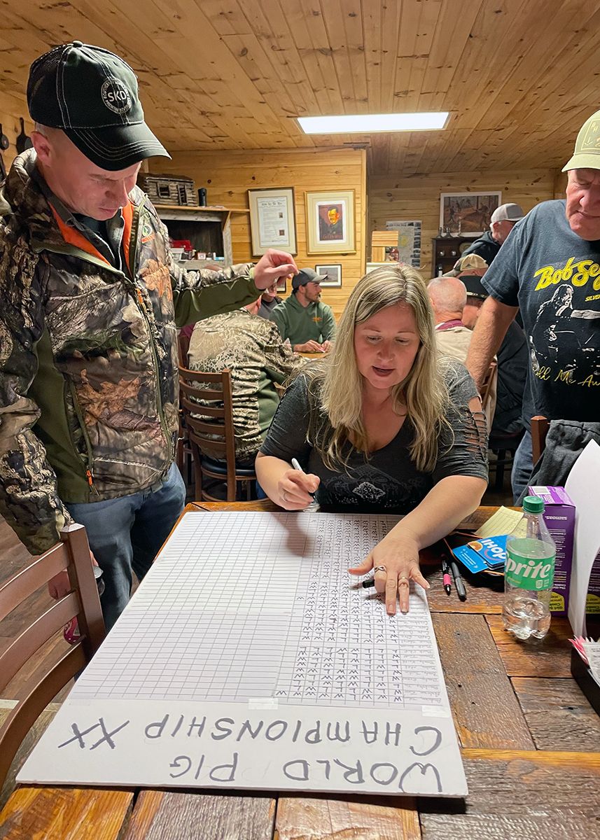 A woman, seated, marks up a large white grid with a handwritten title: World Pig Championships. Two men on either side of her look at the scores.