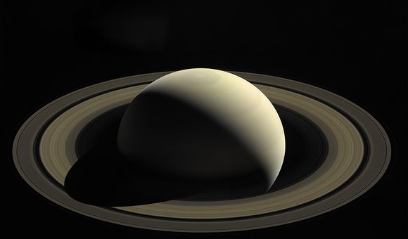 The smooth surface of a round planet is partially illuminated by the sun; the planet's other half is in shadow. The planet is surrounded by thousands of rings.