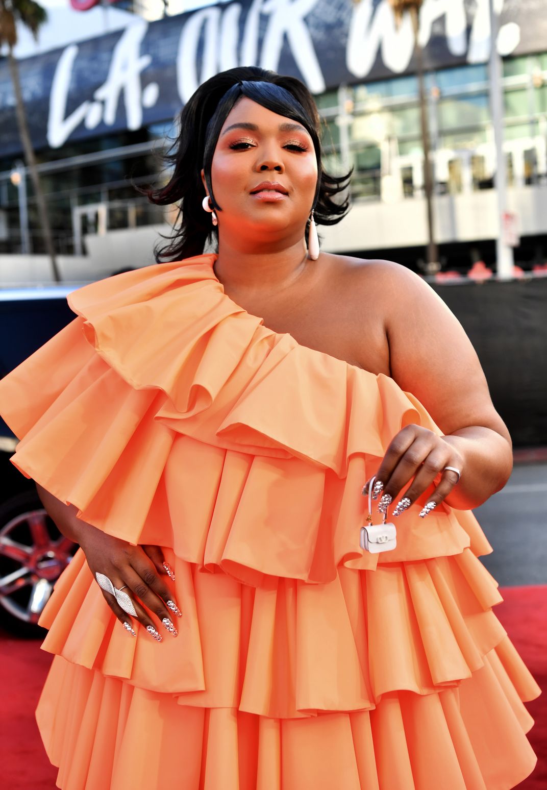 Lizzo wearing an orange dress and holding up a small handbag
