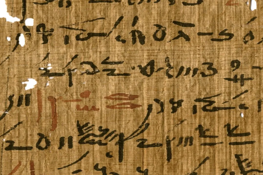A yellow-orange papyrus sheet, with some small holes, with elongated handwritten black hieroglyphic script, with small sections every few lines written in faded red ink
