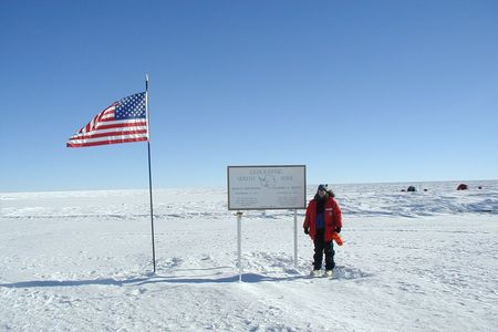 Geolgist Cari Corrigan poses for a picture in the snow next to an American flag and a sign that reads "Geographic South Pole."