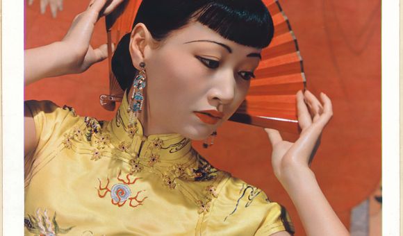 Portrait of Anna May Wong. Wong wears a yellow dress and gazes to the side of the frame. She delicately holds a red folding fan behind her head.