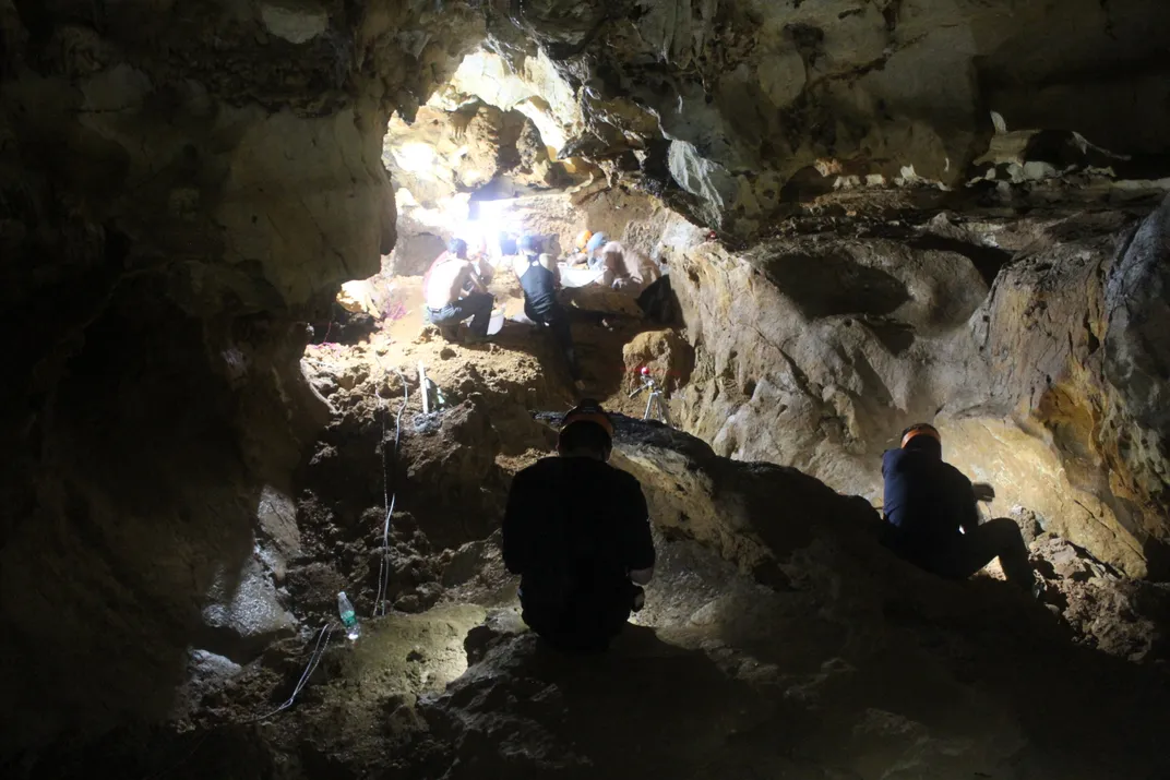 paleontologists working in a cave lit partially by a hole near the top of the image