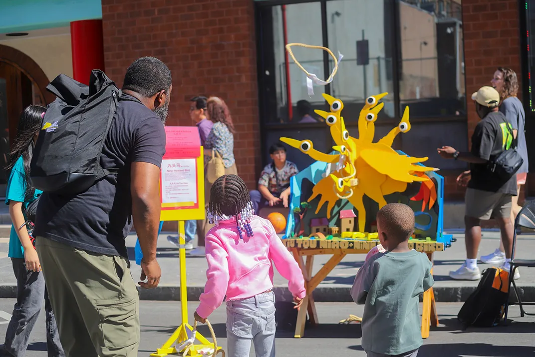 An adult and two children play a ring toss game, tossing a yellow, circular rope toward a yellow nine-headed bird figure. Festival goers pass in the background.
