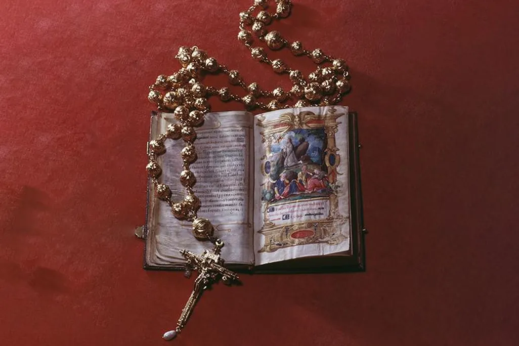 On a red background, an open illuminated Bible with colorful illustrations depicting Jesus kneeling and praying, encircled by a golden rosary with large round beads and a cross decorated with pearls and a golden figure of Jesus crucified on its end