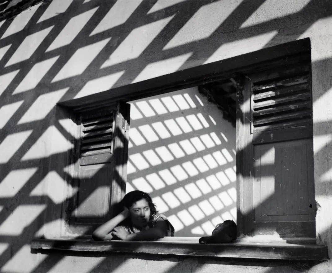 A black and white image of a woman leaning out of a window, with striking black and white grids of shadow covering her face