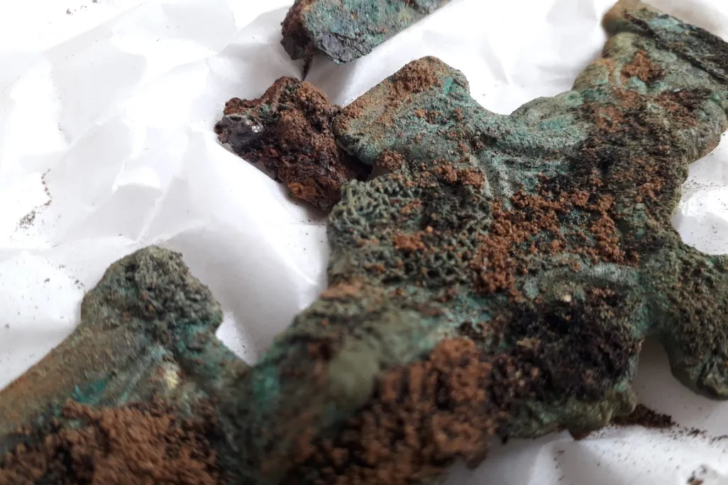 A close-up image of a greenish cross that appears carved and has bits of brown hardened material, textiles, adhering to its surface