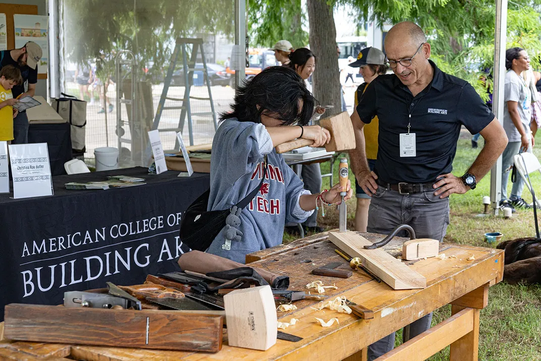 A man stands with hands on his hips, speaking to a person who uses a mallet to chisel a piece of wood on an outdoor work station.