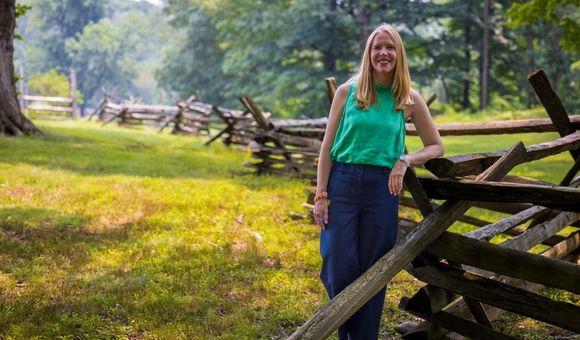 A woman leans on a snaking, split rail fence on a mossy green field.