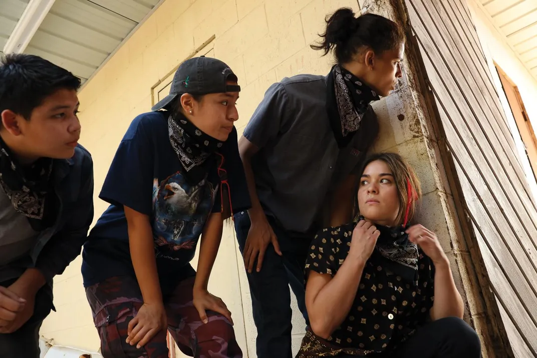 three Native American teenage boys peering around a wall with and one Native teenage girl, who looks back at them