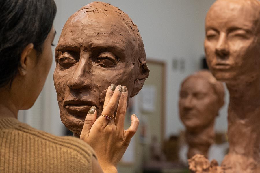 A person sculpts a clay bust. Two additional sculptures appear in the background.