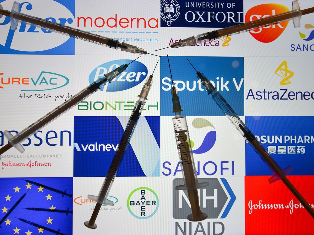 An illustrative image of medical syringes in front of the logos of various companies working on the Covid-19 vaccine, displayed on a screen. 