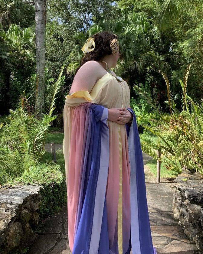 A person poses, facing away, wearing a costume of a flowing sleeveless dress and shawl in pale yellow, pink, and dark blue.